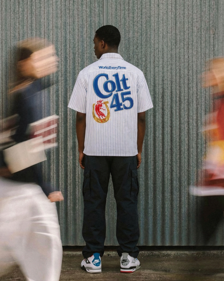 Person wearing a white shirt with 'Colt 45' branding in front of a corrugated metal wall.