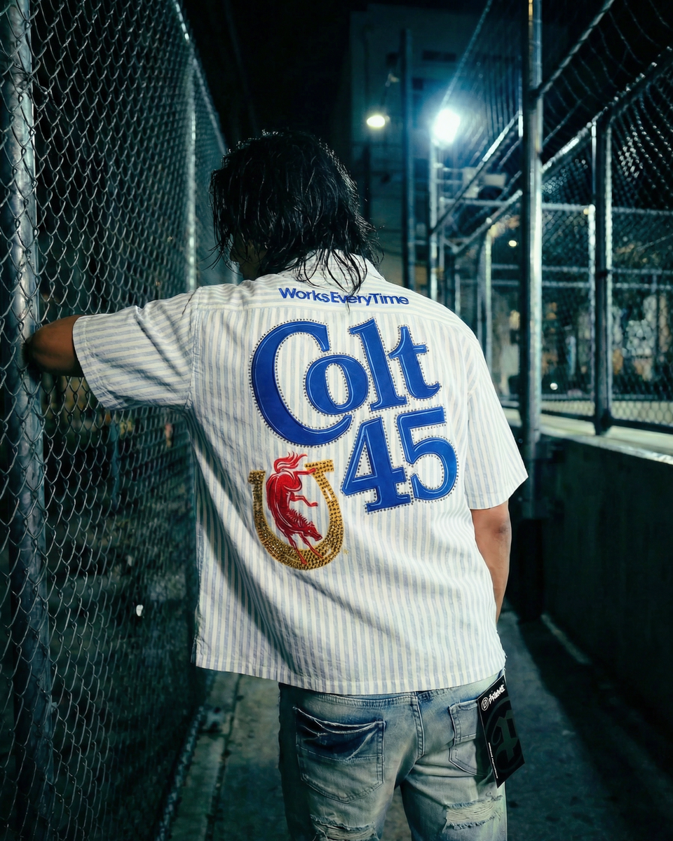 Person wearing a 'Colt 45' shirt leaning against a chain-link fence at night.