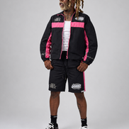 A man modeling a black and pink DUAA track jacket and matching shorts with white graphic details, styled with a white t-shirt, black socks, and black sneakers against a light gray studio background.