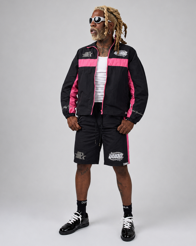 A man modeling a black and pink DUAA track jacket and matching shorts with white graphic details, styled with a white t-shirt, black socks, and black sneakers against a light gray studio background.