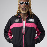 A man with blonde dreadlocks and white sunglasses wearing a black and pink track jacket with 'DUAA' branding, posed against a solid gray studio background.