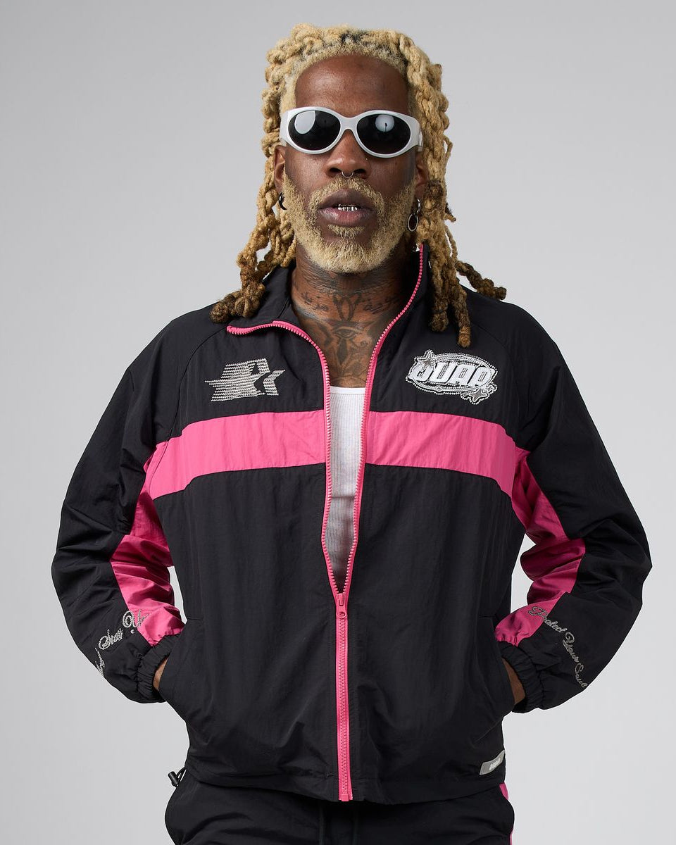 A man with blonde dreadlocks and white sunglasses wearing a black and pink track jacket with 'DUAA' branding, posed against a solid gray studio background.