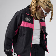 A man wearing a black and pink windbreaker jacket, black pants, a white tank top, and white sunglasses, photographed against a light gray studio background.