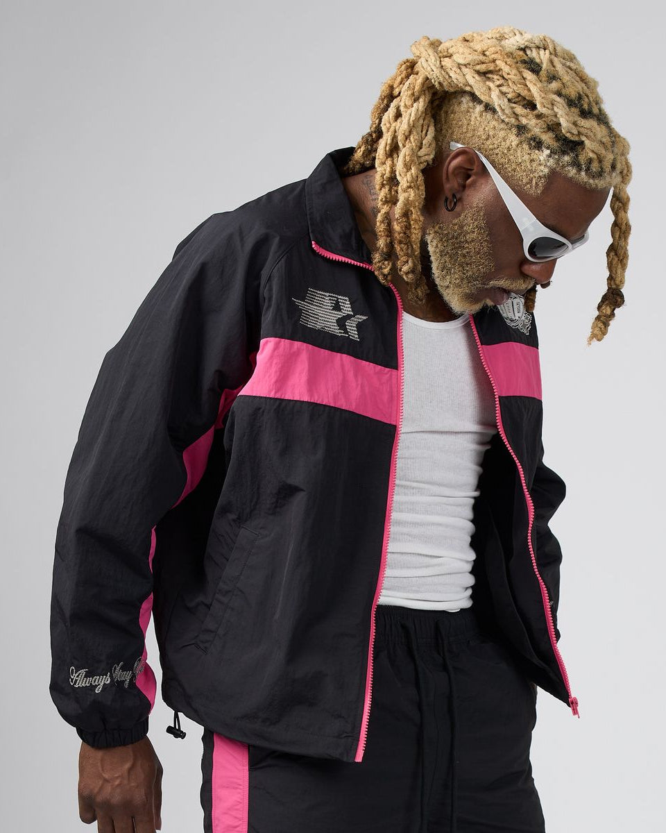 A man wearing a black and pink windbreaker jacket, black pants, a white tank top, and white sunglasses, photographed against a light gray studio background.
