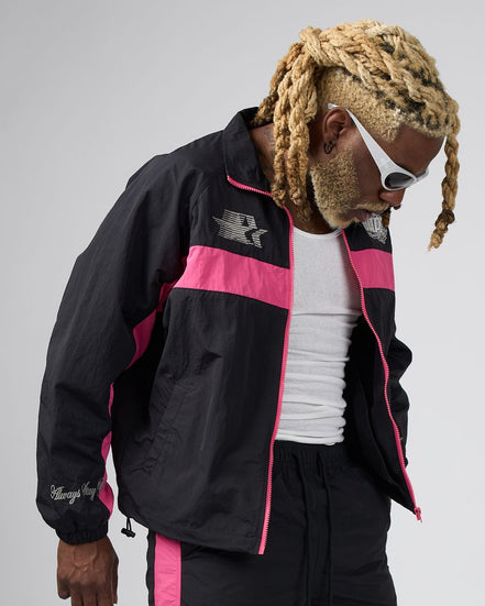 A man wearing a black and pink windbreaker jacket, black pants, a white tank top, and white sunglasses, photographed against a light gray studio background.