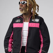 A man wearing a black and pink DUAA track jacket, styled with a white tank top, black pants, and white sunglasses, set against a plain gray studio background.