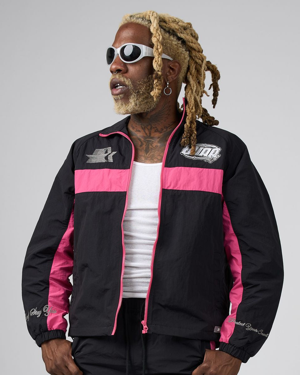 A man wearing a black and pink DUAA track jacket, styled with a white tank top, black pants, and white sunglasses, set against a plain gray studio background.