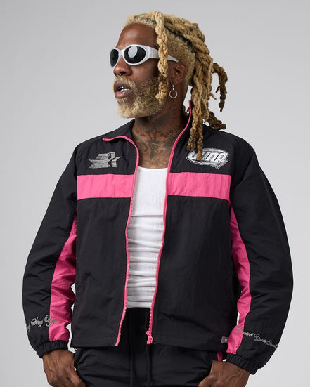 A man wearing a black and pink DUAA track jacket, styled with a white tank top, black pants, and white sunglasses, set against a plain gray studio background.