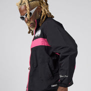 A man with blonde dreadlocks and white sunglasses models a black windbreaker jacket with bright pink color blocking and white script branding on the sleeve, set against a plain light gray background.