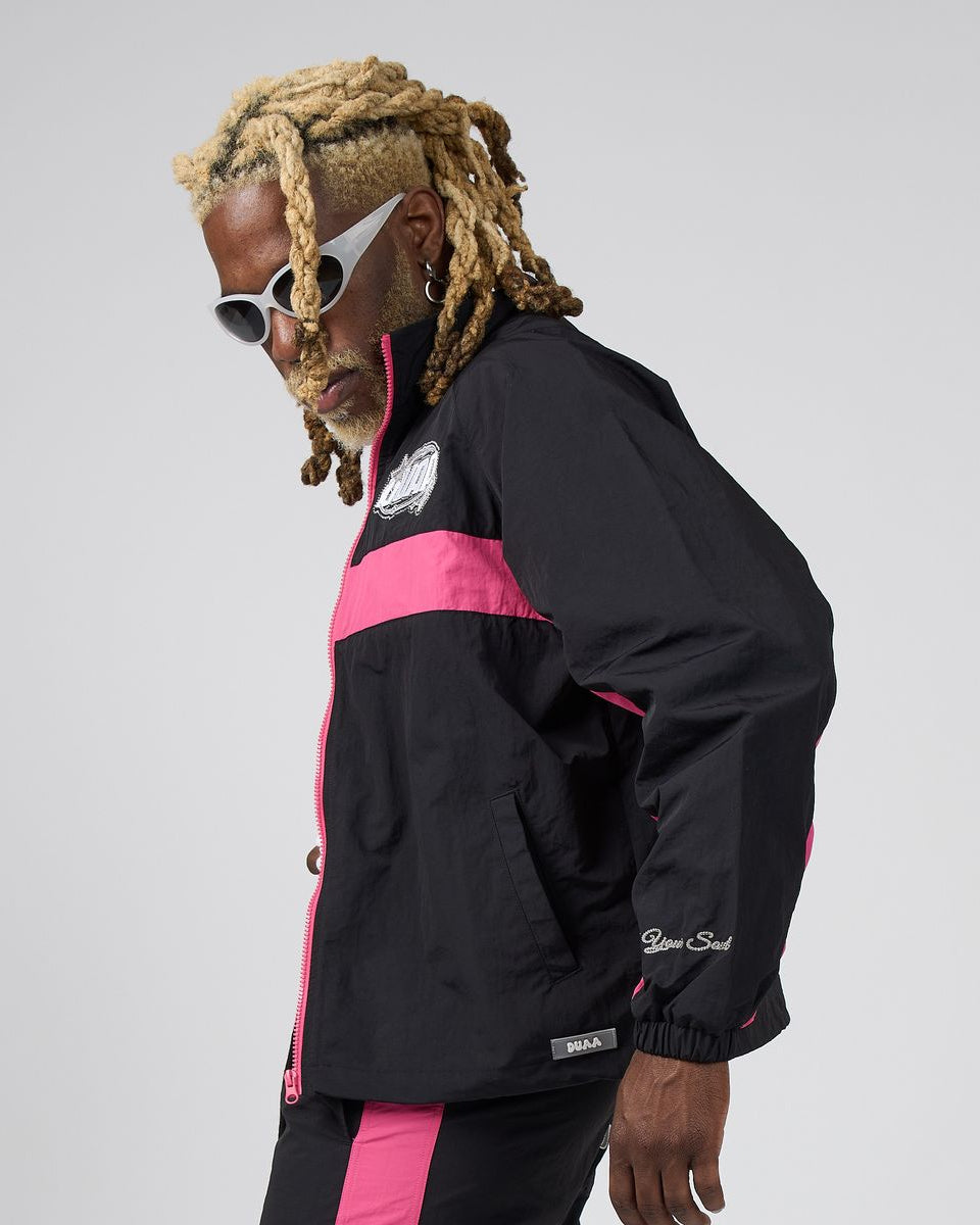 A man with blonde dreadlocks and white sunglasses models a black windbreaker jacket with bright pink color blocking and white script branding on the sleeve, set against a plain light gray background.