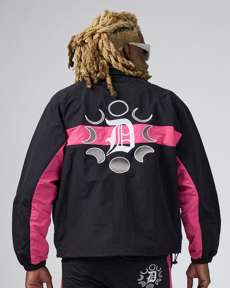 A man viewed from behind wearing a black windbreaker jacket with bright pink side panels and a large white graphic logo on the back, paired with black pants against a light gray studio background.
