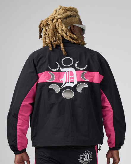 A man viewed from behind wearing a black windbreaker jacket with bright pink side panels and a large white graphic logo on the back, paired with black pants against a light gray studio background.