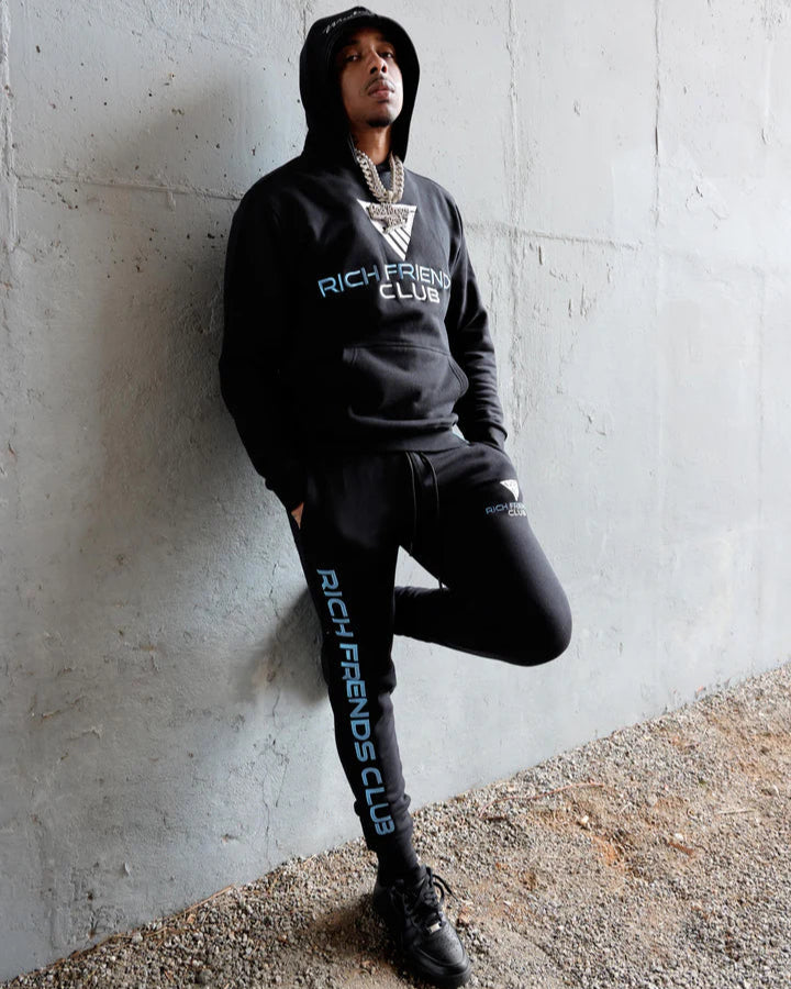 Person wearing a black hoodie and pants with 'Rich Friends Club' branding against a concrete wall.