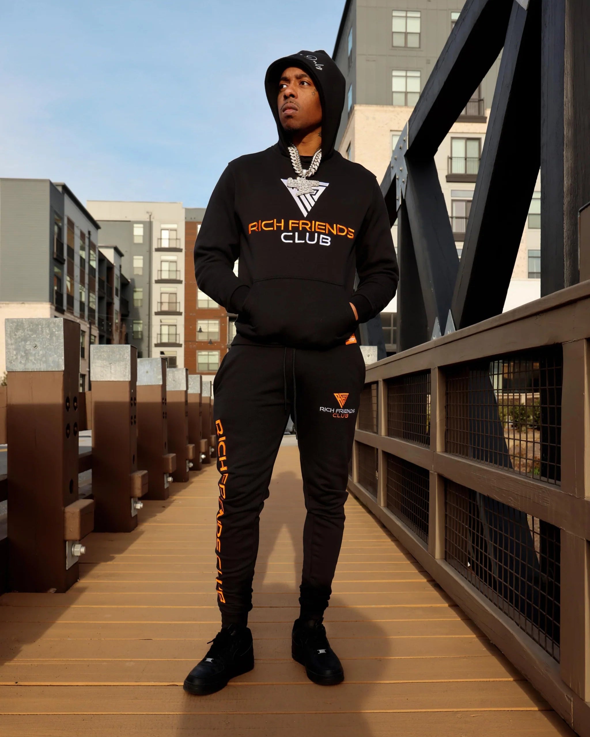 Person wearing a black hoodie and pants with 'Rich Friends Club' branding on a wooden bridge.