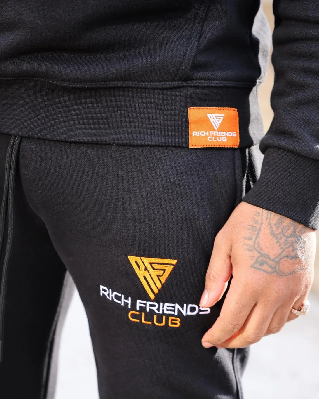 Person wearing black tracksuit with 'Rich Friends Club' logo on a light gray background