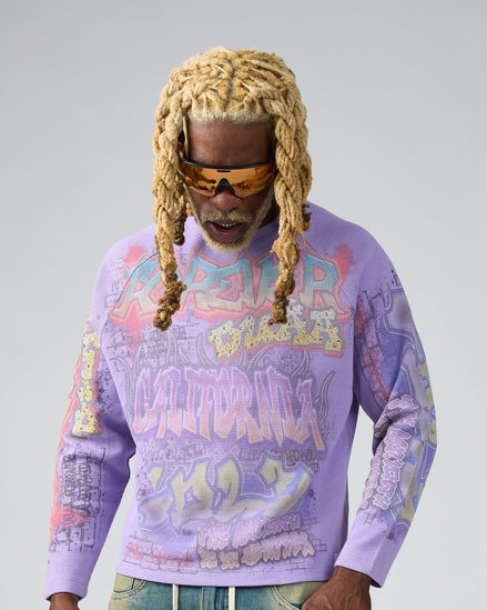 Man wearing a colorful graphic sweatshirt with sunglasses and braided hair on a plain background