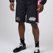 A model wearing black Puma shorts with pink side stripes, a matching black and pink jacket, black socks, and black patent leather sneakers, shown from the waist down on a light gray studio background.