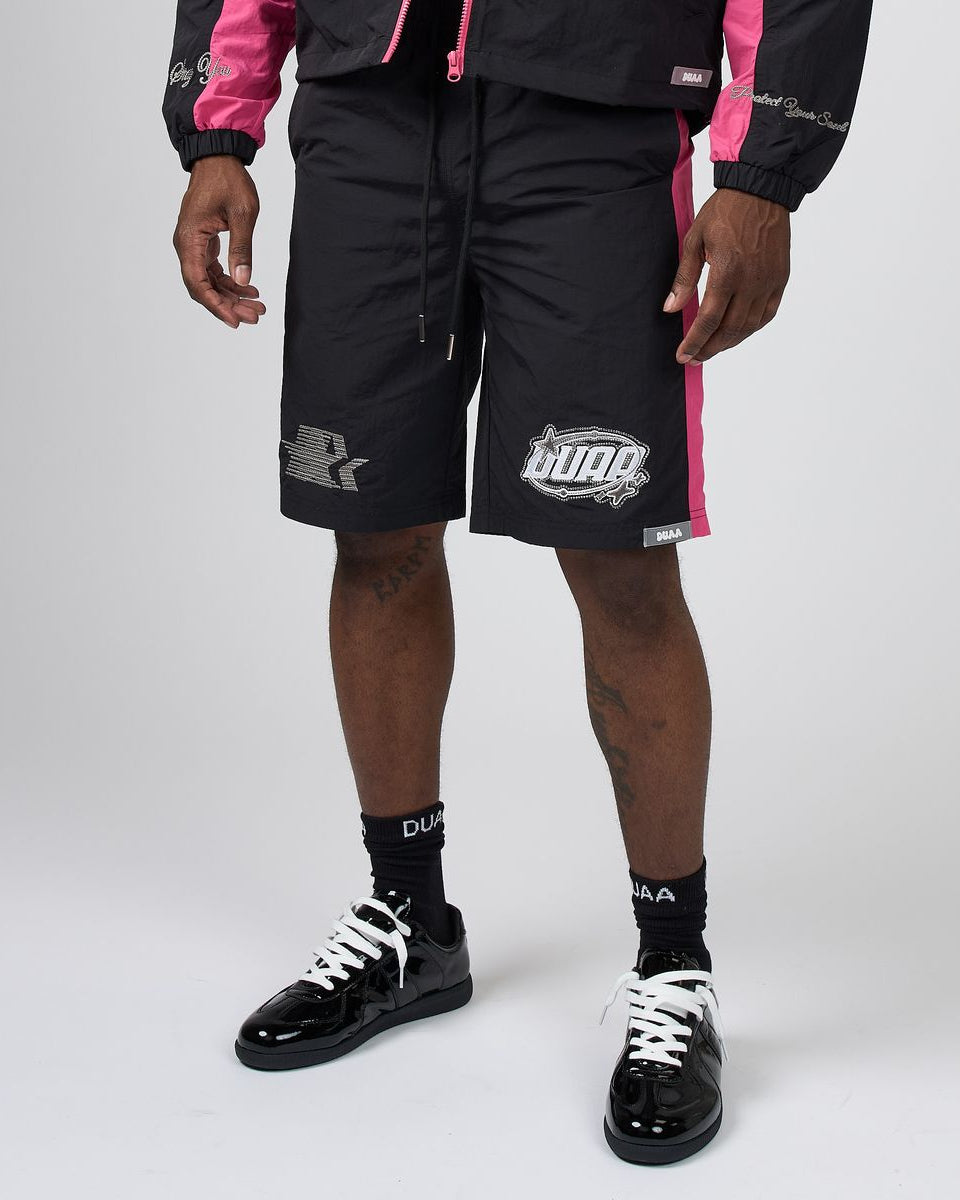 A model wearing black Puma shorts with pink side stripes, a matching black and pink jacket, black socks, and black patent leather sneakers, shown from the waist down on a light gray studio background.