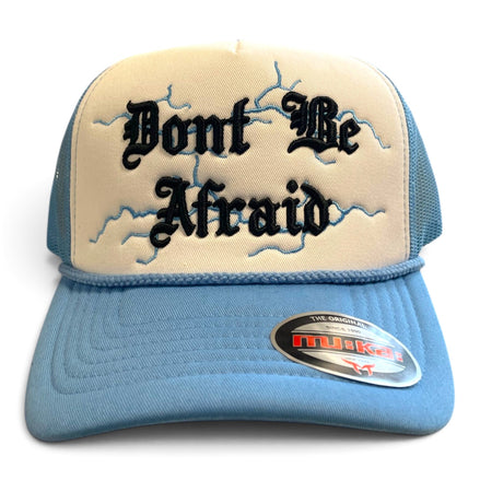 A light blue and cream trucker hat featuring the embroidered text 'Don't Be Afraid' in black Gothic lettering over a blue lightning-bolt design on the front panel. The hat has a light blue curved brim with a visible 'Mueller' brand sticker and cream mesh back panels.