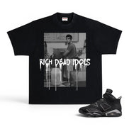 A black short-sleeve graphic T-shirt displayed flat against a white background, paired with a black and gray sneaker. The shirt features a black-and-white photo print of a man in a room with the text 'RICH DEAD IDOLS' in a dripping white font across the front.