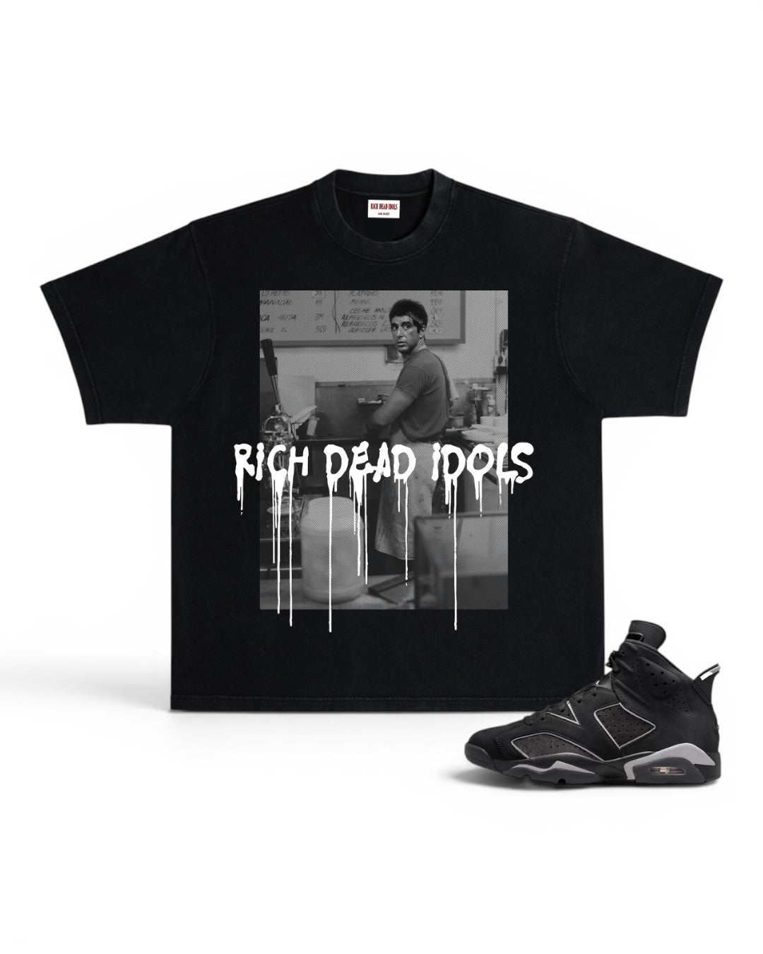 A black short-sleeve graphic T-shirt displayed flat against a white background, paired with a black and gray sneaker. The shirt features a black-and-white photo print of a man in a room with the text 'RICH DEAD IDOLS' in a dripping white font across the front.