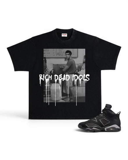 A black short-sleeve graphic T-shirt displayed flat against a white background, paired with a black and gray sneaker. The shirt features a black-and-white photo print of a man in a room with the text 'RICH DEAD IDOLS' in a dripping white font across the front.