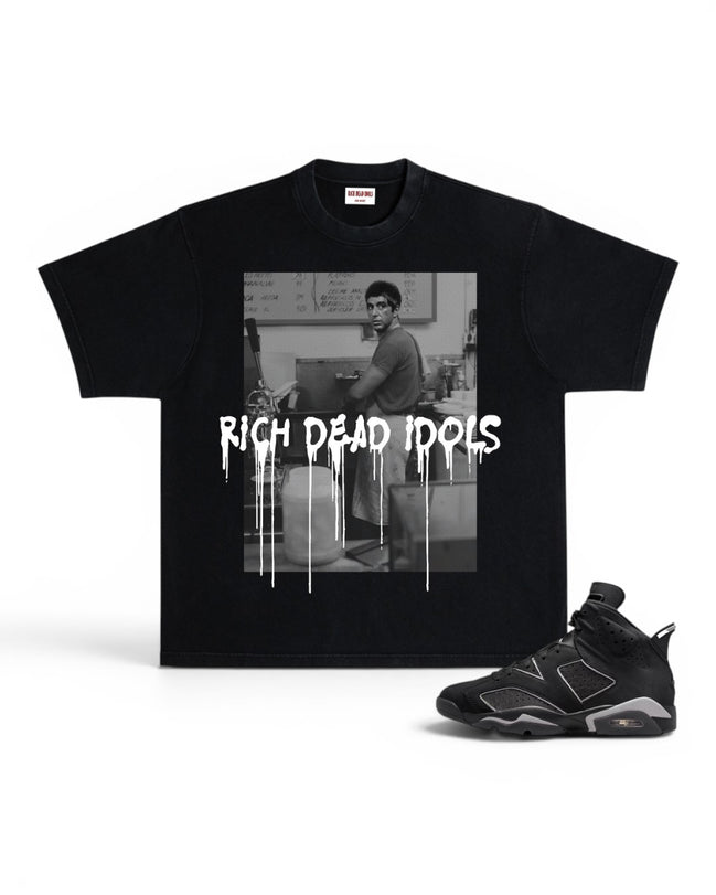 A black short-sleeve graphic T-shirt displayed flat against a white background, paired with a black and gray sneaker. The shirt features a black-and-white photo print of a man in a room with the text 'RICH DEAD IDOLS' in a dripping white font across the front.