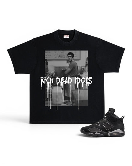 A black short-sleeve graphic T-shirt displayed flat against a white background, paired with a black and gray sneaker. The shirt features a black-and-white photo print of a man in a room with the text 'RICH DEAD IDOLS' in a dripping white font across the front.