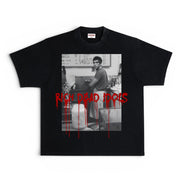 A black short-sleeve graphic t-shirt featuring a black-and-white photo print of a man in a kitchen, overlaid with the words 'RICH DEAD IDOLS' in dripping red text.