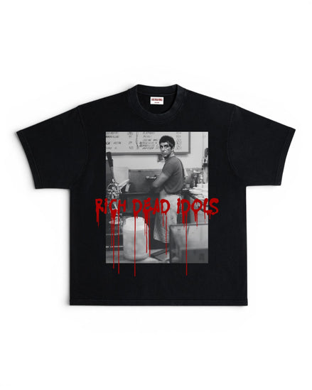 A black short-sleeve graphic t-shirt featuring a black-and-white photo print of a man in a kitchen, overlaid with the words 'RICH DEAD IDOLS' in dripping red text.