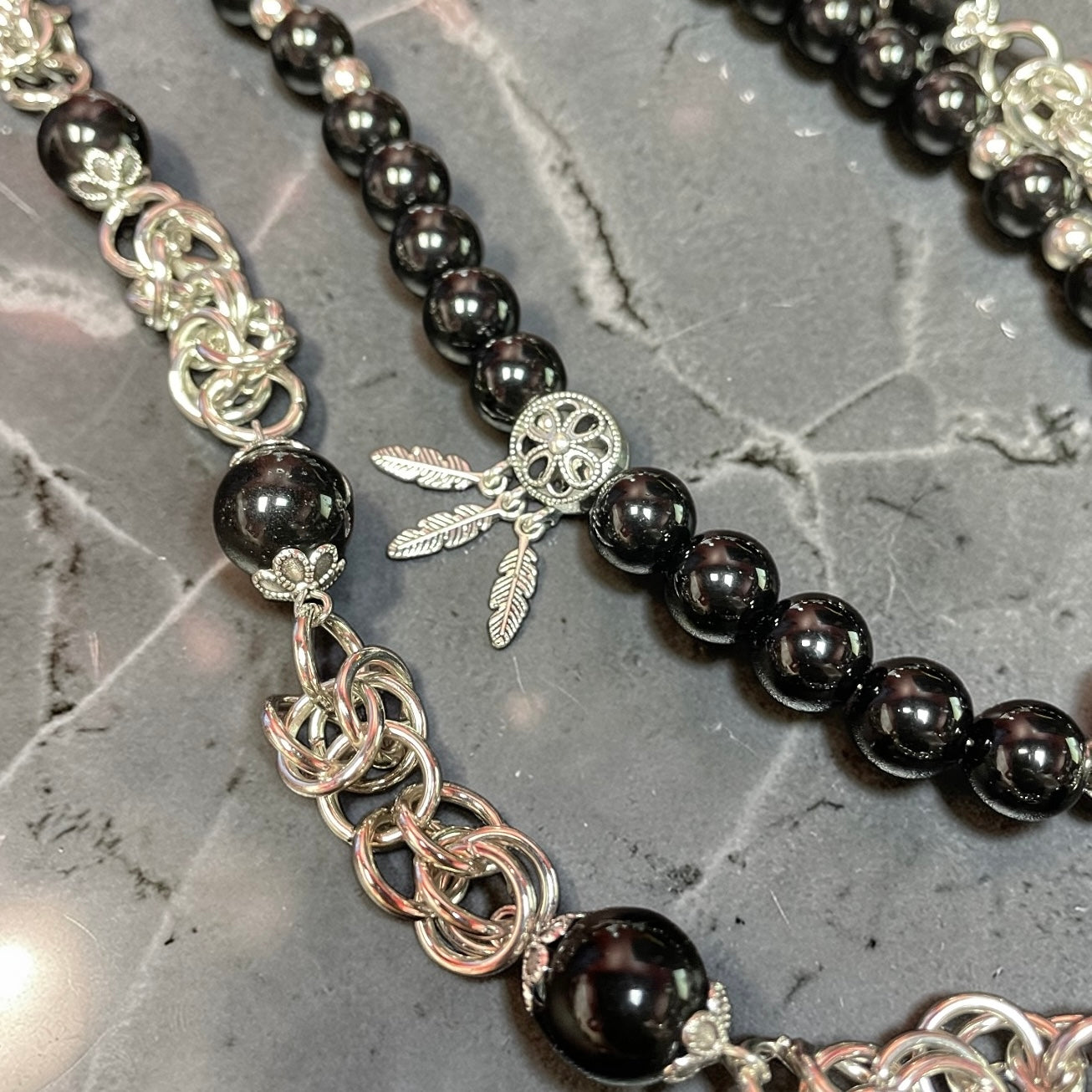 Close-up of a black beaded necklace with silver chain and feather charm on a gray stone surface.