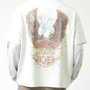 A person shown from the back wearing a light gray oversized T-shirt with a large distressed eagle graphic and a shield-style patch design in brown, red, yellow, and blue tones. The shirt has long gray sleeves and is paired with tan pants against a plain white studio background.