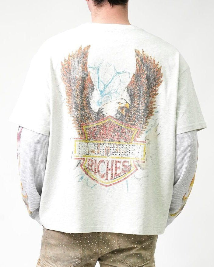 A person shown from the back wearing a light gray oversized T-shirt with a large distressed eagle graphic and a shield-style patch design in brown, red, yellow, and blue tones. The shirt has long gray sleeves and is paired with tan pants against a plain white studio background.