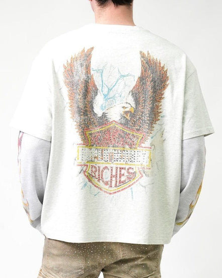 A person shown from the back wearing a light gray oversized T-shirt with a large distressed eagle graphic and a shield-style patch design in brown, red, yellow, and blue tones. The shirt has long gray sleeves and is paired with tan pants against a plain white studio background.