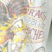 A close-up of a graphic t-shirt featuring a distressed, vintage-style illustration of a Native American figure with a feathered headdress and a bicycle, with the word 'RACE' printed in red on a white sign.
