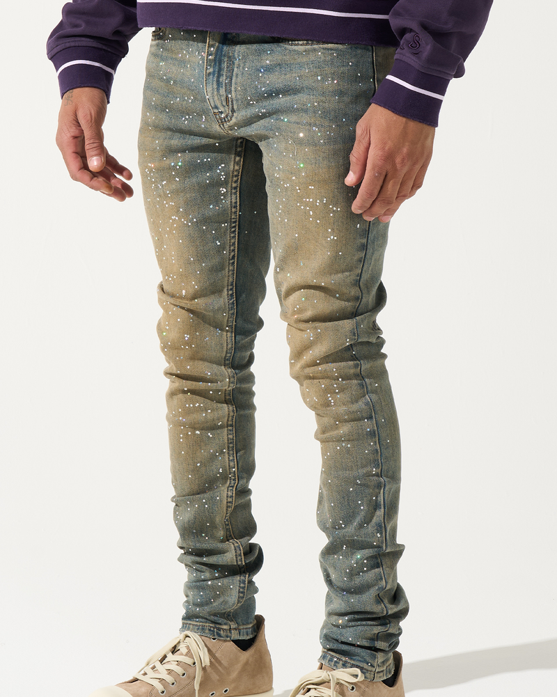 Person wearing blue jeans with white speckles and beige sneakers on a white background