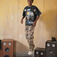 Person wearing a black graphic t-shirt and camouflage pants standing between two speakers against a textured wall.