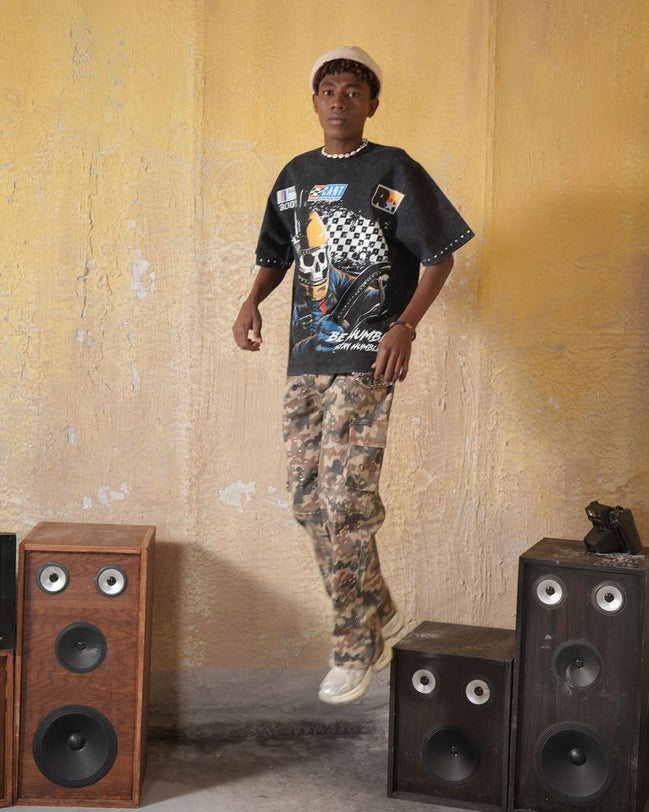 Person wearing a black graphic t-shirt and camouflage pants standing between two speakers against a textured wall.