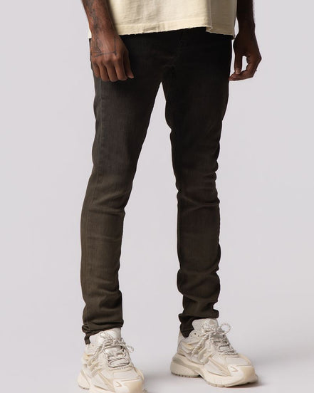 A model from the waist down wearing slim-fit dark brown jeans and chunky off-white sneakers, standing against a light gray studio background.