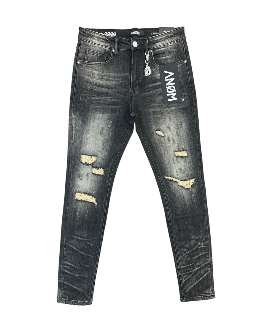 Distressed black denim jeans with ripped details showing beige lining, featuring a white vertical 'ANOM' logo on the right thigh, displayed flat against a white background.