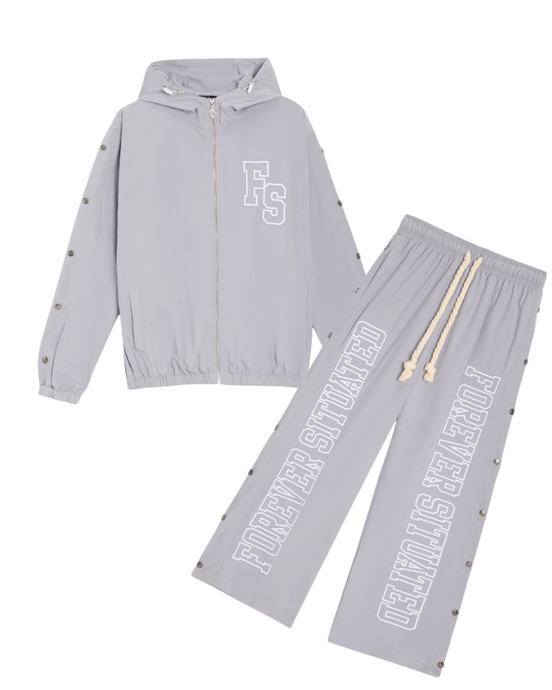 A two-piece light gray tracksuit set featuring a zip-up hooded jacket and matching drawstring pants. The jacket has a white 'FS' monogram on the chest, and the pants feature 'FOREVER SITUATED' printed vertically in white on one leg.