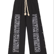 Black wide-leg sweatpants with white 'FOREVER SITUATED' text printed down both legs, featuring a cream-colored rope drawstring and small metallic rivet details along the outer seams.