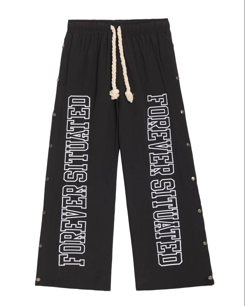 Black wide-leg sweatpants with white 'FOREVER SITUATED' text printed down both legs, featuring a cream-colored rope drawstring and small metallic rivet details along the outer seams.