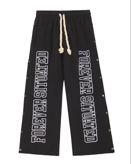 Black wide-leg sweatpants with white 'FOREVER SITUATED' text printed down both legs, featuring a cream-colored rope drawstring and small metallic rivet details along the outer seams.