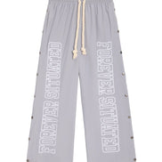 Light gray wide-leg sweatpants with 'FOREVER SITUATED' printed vertically in white on both legs, featuring a cream rope drawstring and silver metal studs along the sides.