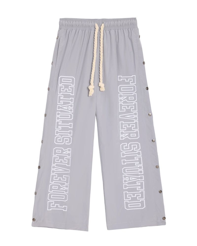 Light gray wide-leg sweatpants with 'FOREVER SITUATED' printed vertically in white on both legs, featuring a cream rope drawstring and silver metal studs along the sides.