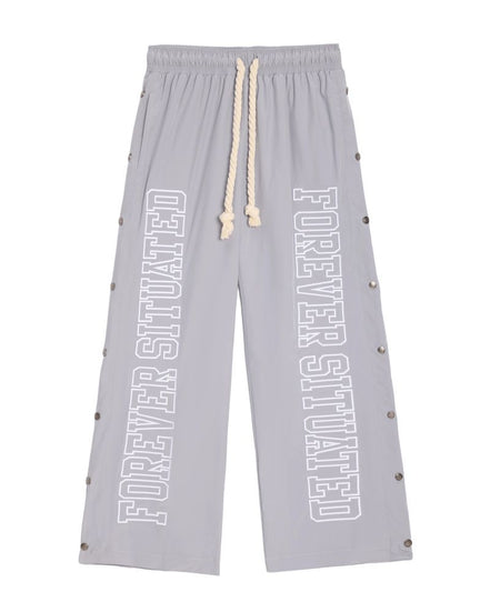 Light gray wide-leg sweatpants with 'FOREVER SITUATED' printed vertically in white on both legs, featuring a cream rope drawstring and silver metal studs along the sides.