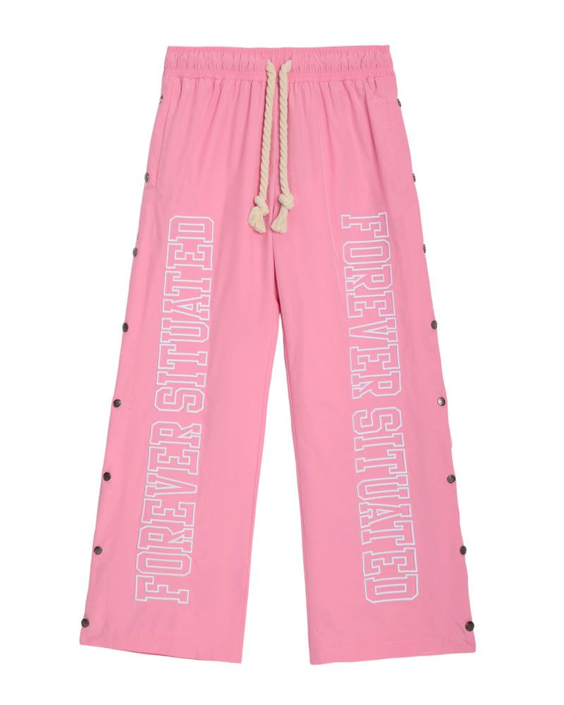Pink wide-leg sweatpants with 'FOREVER SITUATED' printed vertically in white outline text on both legs, featuring a cream rope drawstring and black snap button details along the sides.