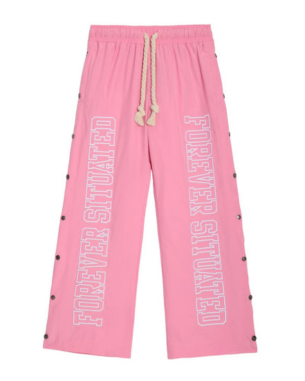 Pink wide-leg sweatpants with 'FOREVER SITUATED' printed vertically in white outline text on both legs, featuring a cream rope drawstring and black snap button details along the sides.