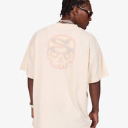 A man shown from the back wearing an oversized cream-colored t-shirt with a large, faded pink and orange skull graphic on the back, paired with light-wash jeans.